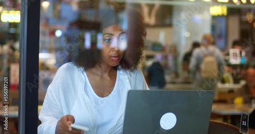 Multi-ethnic woman freelance entrepreneur using mobile phone and laptop, working remotely, planning new business project, sitting in a coffee shop, with blurred unrecognizable customers in background photo