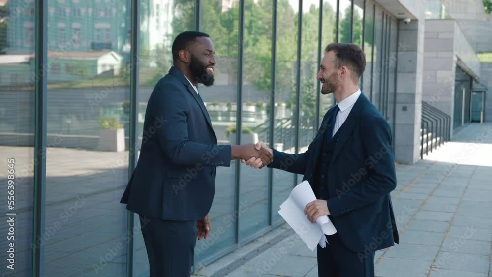 Two business men shake hands when meeting, agree to deal or say hello. Portrait of handshake of two smiling business men making a deal. Office building on the background. Business district. Two business men shake hands when meeting, agree to deal or say hello. Portrait of handshake of two smiling business men making a deal. Office building on the background. Business district.