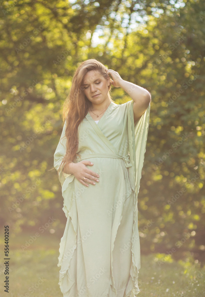 Young pregnant woman stands in park,looks at belly, green trees on background. Brown haired female wears dress.Magic happy pregnancy. Childbirth preparation, emotional connection with baby.Vertical Young pregnant woman stands in park,looks at belly, green trees on background. Brown haired female wears dress.Magic happy pregnancy. Childbirth preparation, emotional connection with baby.Vertical