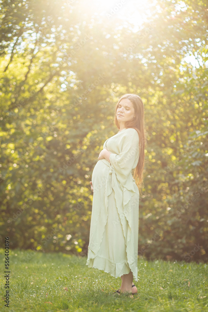 Young pregnant woman holds big belly, green trees, sunlight on background. Brown haired female wear dress. Magic happy pregnancy. Child delivery preparations,emotional connection with baby. Vertical