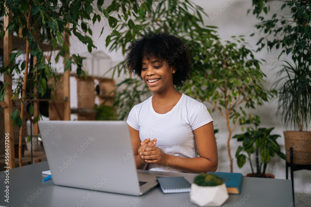 Portrait of cheerful African-American young woman talking during online remote video conference call on laptop, sitting at desk in light home office room with modern green biophilic interior design. Portrait of cheerful African-American young woman talking during online remote video conference call on laptop, sitting at desk in light home office room with modern green biophilic interior design.