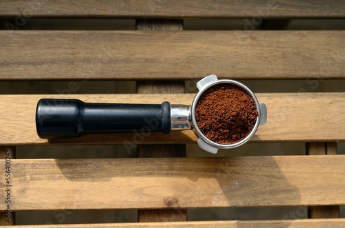 A metal horn of a professional machine coffee filled with brown powder of ground coffee beans stands on a wooden table lit by sunlight, top view.
