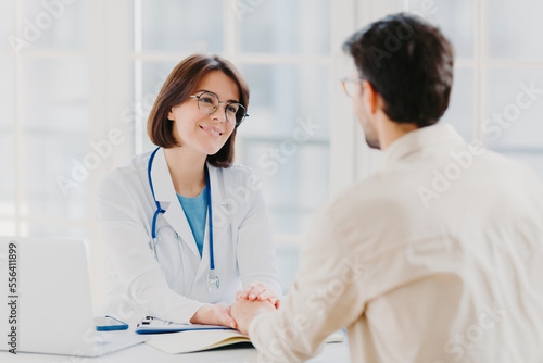 Confident female doctor holds hands of ill patient  persuades everything will be alright  dressed in white medical gown  gives advice  pose in hospial office. Consultation and diagnosis concept