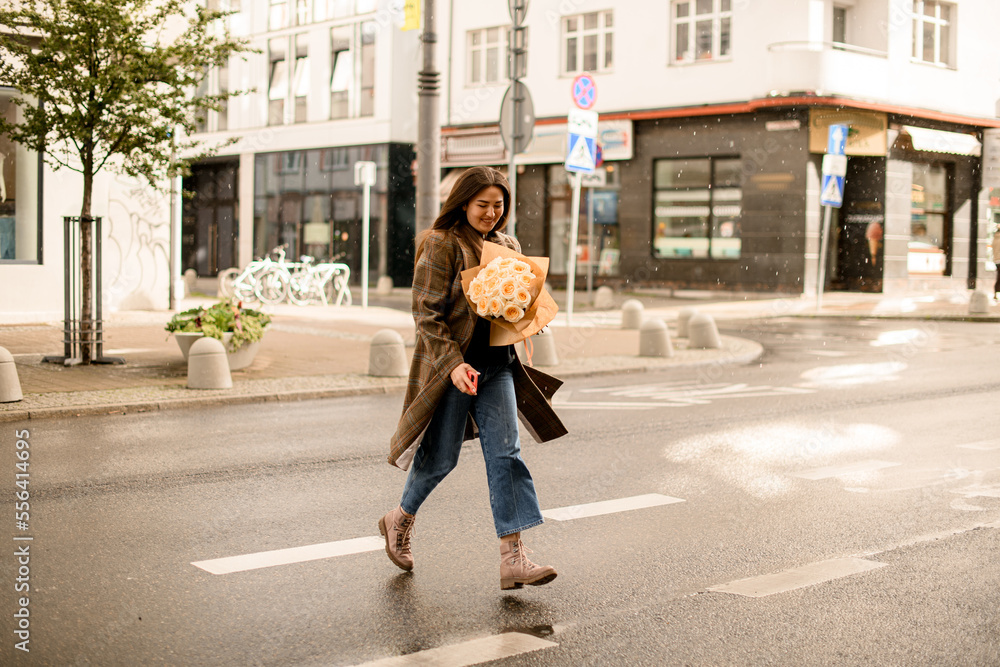 smiling woman in coat with bouquet of cream roses in her hands walking at street
