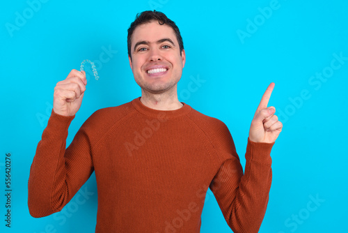 Young handsome Caucasian dark haired man standing over blue studio background holding an invisible aligner braces and pointing aside at empty space. 