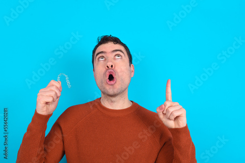 Young handsome Caucasian dark haired man standing over blue studio background holding an invisible aligner braces and pointing with finger up at copy space. 