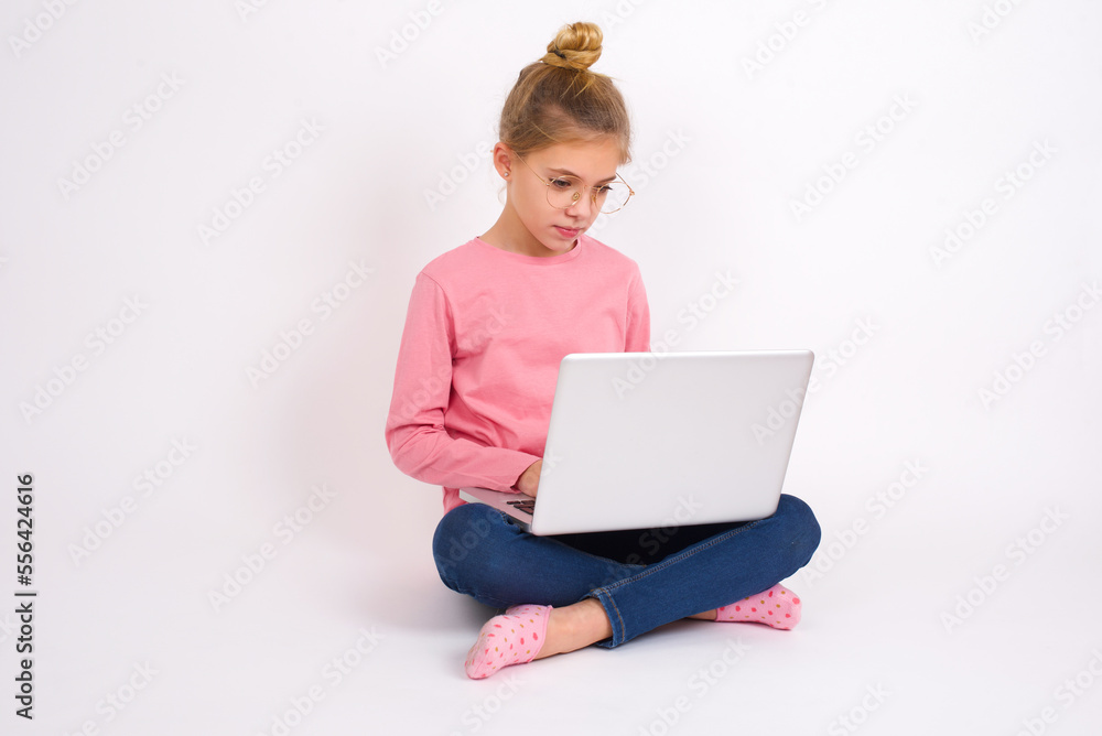 Young beautiful teen girl sitting over white studio background holding a laptop and doing her homework essay. Modern technologies and education concept. Young beautiful teen girl sitting over white studio background holding a laptop and doing her homework essay. Modern technologies and education concept.