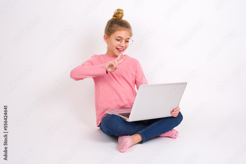 Young beautiful teen girl sitting over white studio background holding a laptop, speaking with classmates via conference call and making v sign with fingers. Young beautiful teen girl sitting over white studio background holding a laptop, speaking with classmates via conference call and making v sign with fingers.