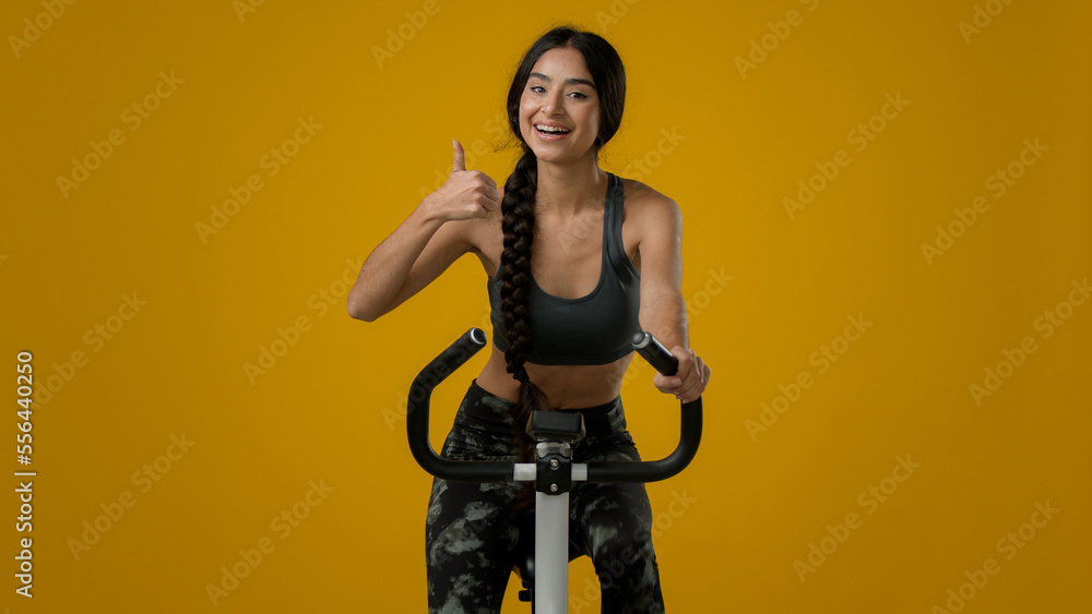 Obraz premium Indian ethnic smiling sporty woman female lady athletic girl cyclist biking flexing cycling riding on exercise bike sport equipment fitness workout cardio ride in yellow studio thumb up recommend