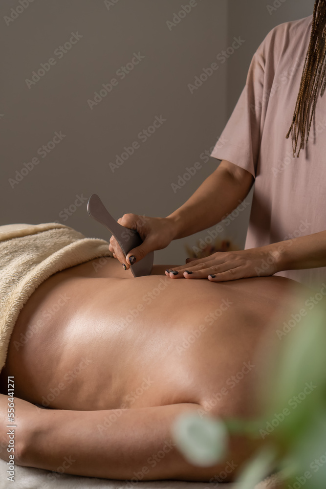 Obraz premium Close-up of a masseur does a back massage with a metal massage tool to a woman lying on a couch in a cozy office. Health concept, body care, skin care, wellness. Vertical photo