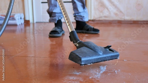 vacuum cleaner pipe with a black plastic brush sucks gray dust from a brown wooden floor and men's feet on a blurred background, cleaning an old dusty room