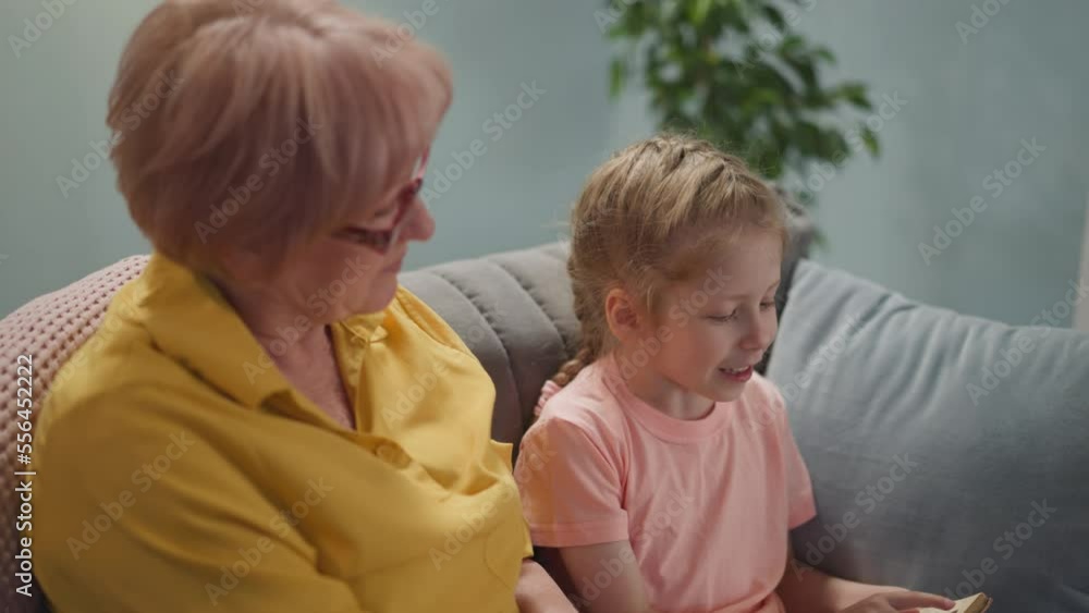 Happy senior lady sits near little girl reading book in living room slow motion. Grandparent helps preschooler child to do homework. Home education Happy senior lady sits near little girl reading book in living room slow motion. Grandparent helps preschooler child to do homework. Home education