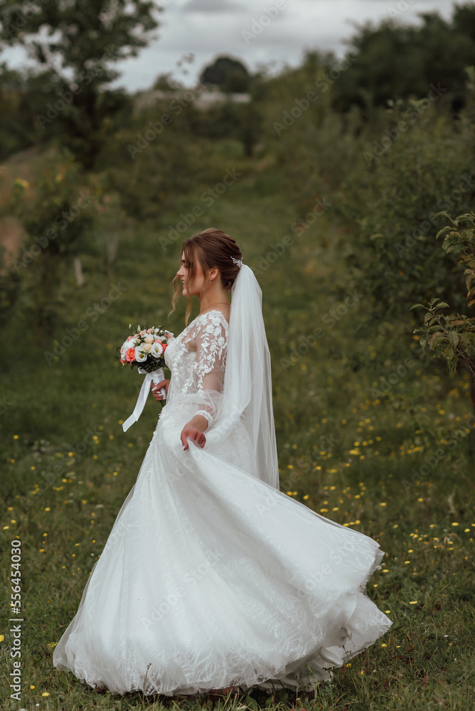 a young beautiful bride poses with a wedding bouquet against the background of nature; the girl is dressed in a white wedding dress; a moment on the wedding day; happy girl expresses her emotions