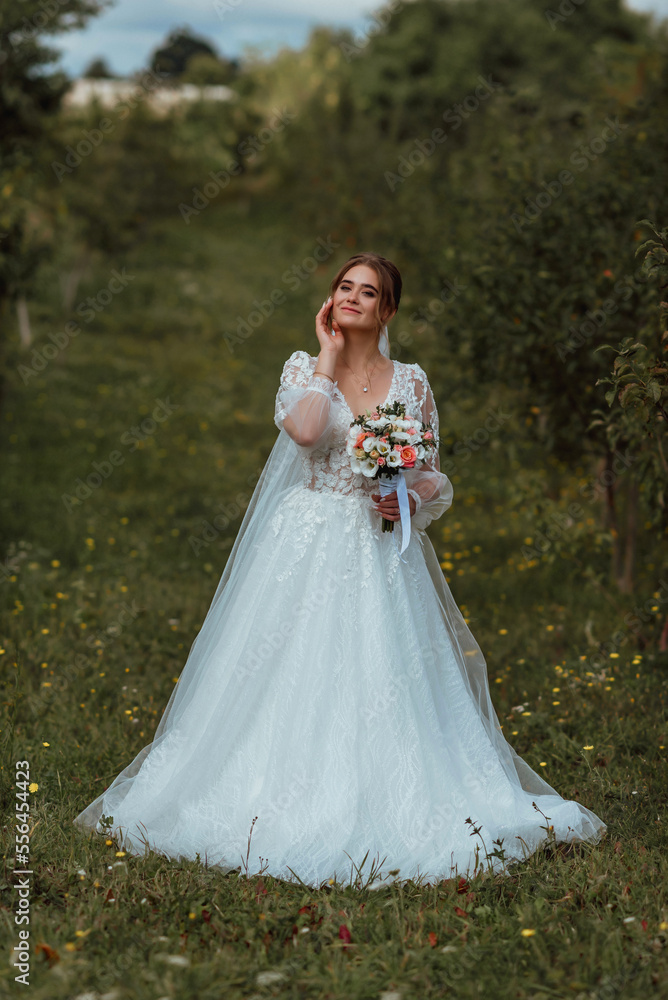 a young beautiful bride poses with a wedding bouquet against the background of nature; the girl is dressed in a white wedding dress; a moment on the wedding day; happy girl expresses her emotions