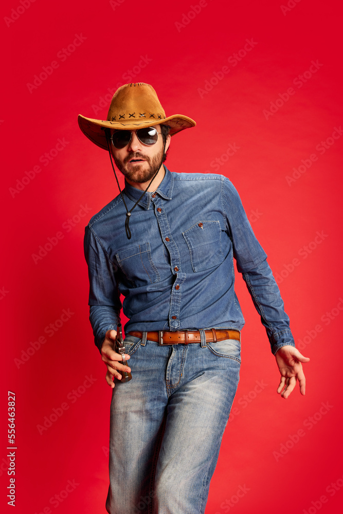 Excited young man in cowboy style outfit having fun, posing isolated over red background. Model in cowboy hat and denim clothes. Fashion, emotions concept Excited young man in cowboy style outfit having fun, posing isolated over red background. Model in cowboy hat and denim clothes. Fashion, emotions concept