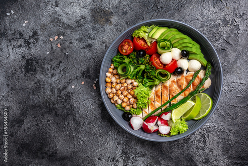 Grilled chicken, avocado, asparagus, chickpeas, broccoli, radish, cucumber, tomatoes, olives, mozzarella buddha bowl on dark background, top view. Delicious balanced food concept
