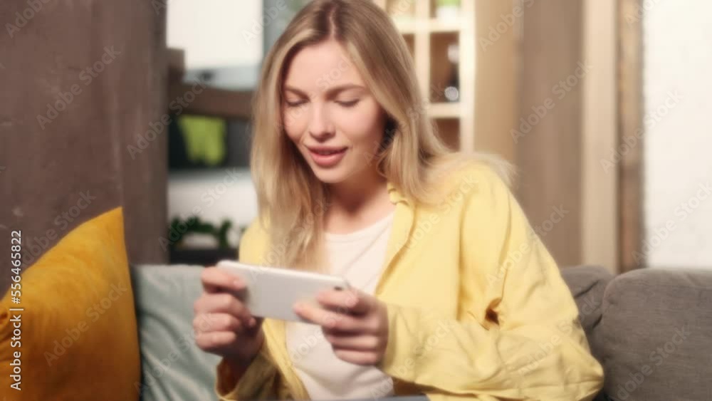 Soft dreamy portrait of charming blond woman gamer sitting on sofa relaxing alone and playing in online video games on smartphone holding in horizontal landscape mode at living room at home Soft dreamy portrait of charming blond woman gamer sitting on sofa relaxing alone and playing in online video games on smartphone holding in horizontal landscape mode at living room at home