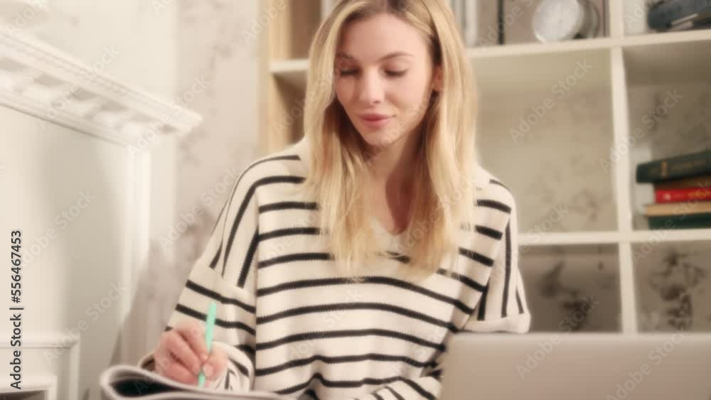 Dreamy soft shot of attractive young blond girl preparing for exam and studying at home Charming young student focused on learning and make notes in her notebook while looking at laptop computer Dreamy soft shot of attractive young blond girl preparing for exam and studying at home Charming young student focused on learning and make notes in her notebook while looking at laptop computer