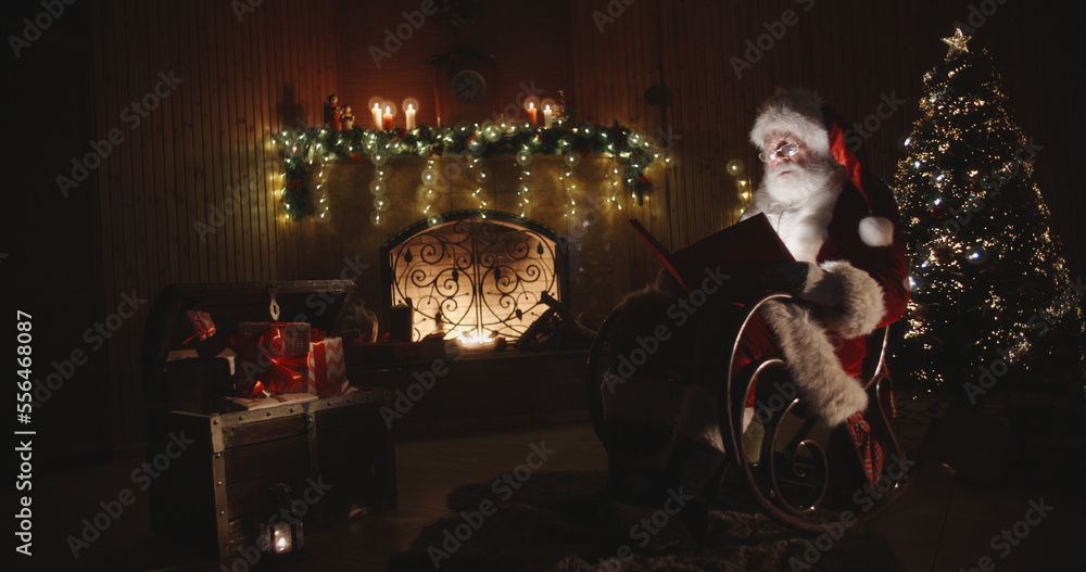 Santa claus sitting on roker in specially decorated room, reading a magical shining book - holidays and celebrations, christmas spirit concept 