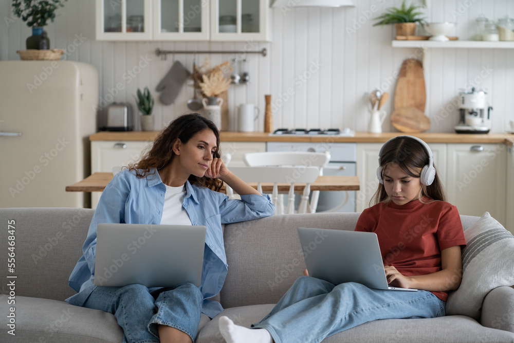 Mom freelancer looking at daughter sitting beside and studying learning remotely. Mother and teen girl kid sitting on sofa with laptops, working online with children. Distance education and parenting Mom freelancer looking at daughter sitting beside and studying learning remotely. Mother and teen girl kid sitting on sofa with laptops, working online with children. Distance education and parenting