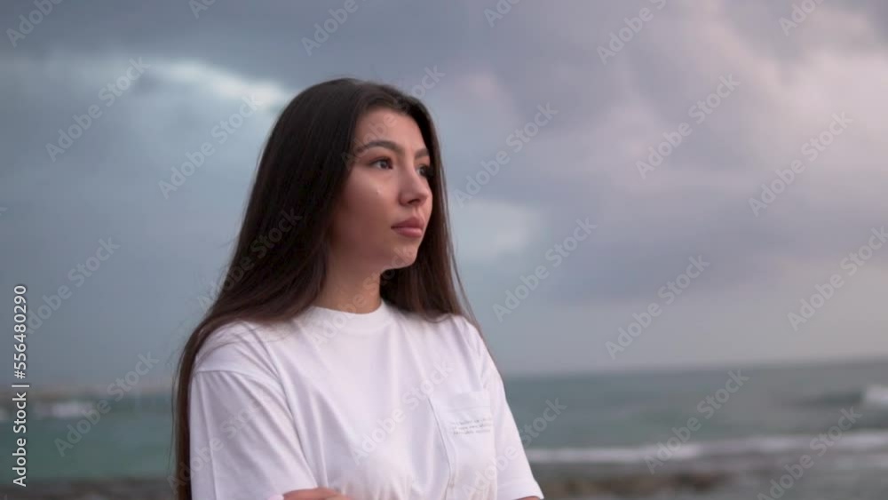 Close up portrait of Young Woman Serious with thick black hair looking at sunset over ocean. Cloudy rainy day, slow shooting. Beautiful woman looks into the distance Close up portrait of Young Woman Serious with thick black hair looking at sunset over ocean. Cloudy rainy day, slow shooting. Beautiful woman looks into the distance