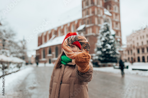 Xmas tourist woman in a bright hat and warm jacket holding baked obwarzanek traditional polish cuisine snack bagel on old city Market square in Krakow. Merry Christmas and Happy Holidays.  photo