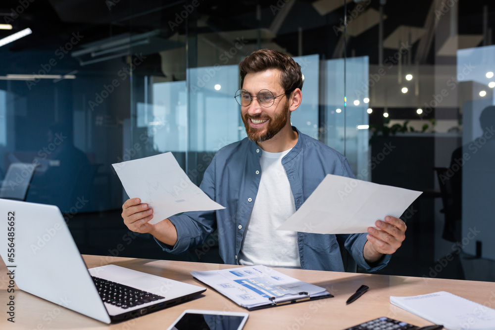 Satisfied with the result financier businessman holds in his hands and examines contracts and reports smiling and happy, man in a casual shirt works inside the office using a laptop in paperwork. Satisfied with the result financier businessman holds in his hands and examines contracts and reports smiling and happy, man in a casual shirt works inside the office using a laptop in paperwork.