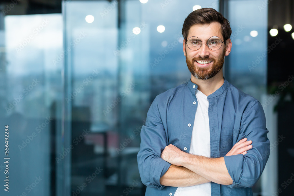 Fototapeta premium Portrait of successful businessman inside modern office, mature man with beard and glasses smiling and looking at camera, boss in casual clothes shirt near window with crossed arms.