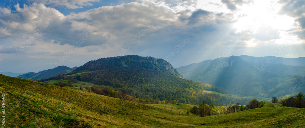 Rural idyllic landscape with grassy fields and rolling hills, sunny afternoon, dramatic sun rays piercing the fluffy clouds. Wonderful springtime landscape in mountains. Nature freshness concept