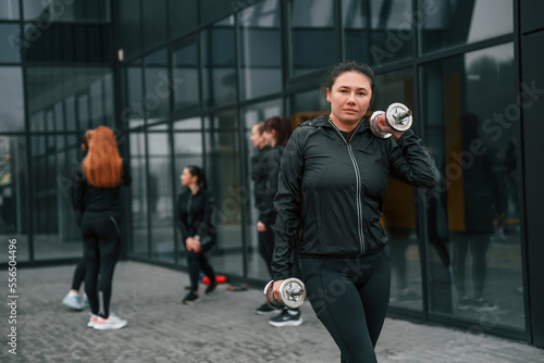 Standing and holding dumbbells in hands. Group of sportive women is outdoors near black building