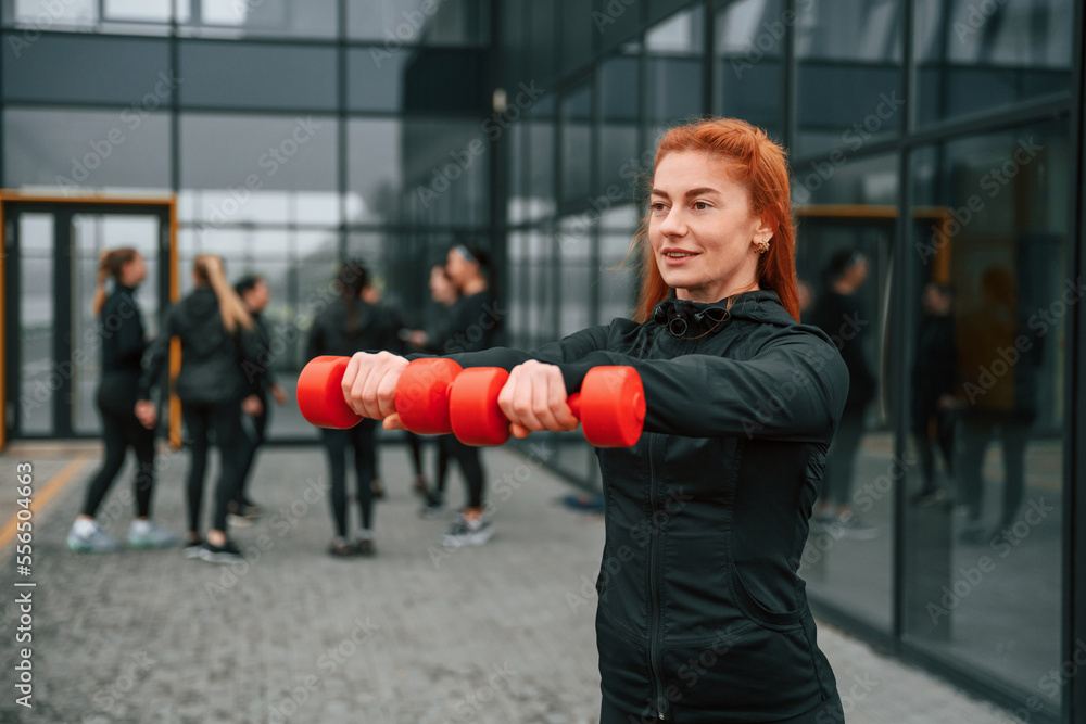 Holding dumbbells. Group of sportive women is outdoors near black building Holding dumbbells. Group of sportive women is outdoors near black building