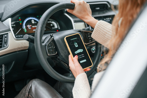 Sitting by steering wheel and holding smartphone that shows charging process. Young woman is in the automobile