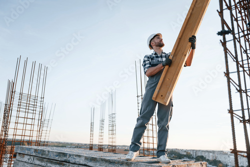 In grey uniform. Holding wooden plank. Man is working on the construction site at daytime