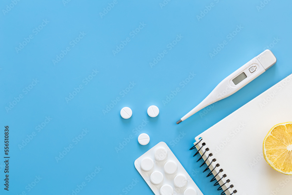 Electronic thermometer, pills and notepad on a blue background, flat lay.