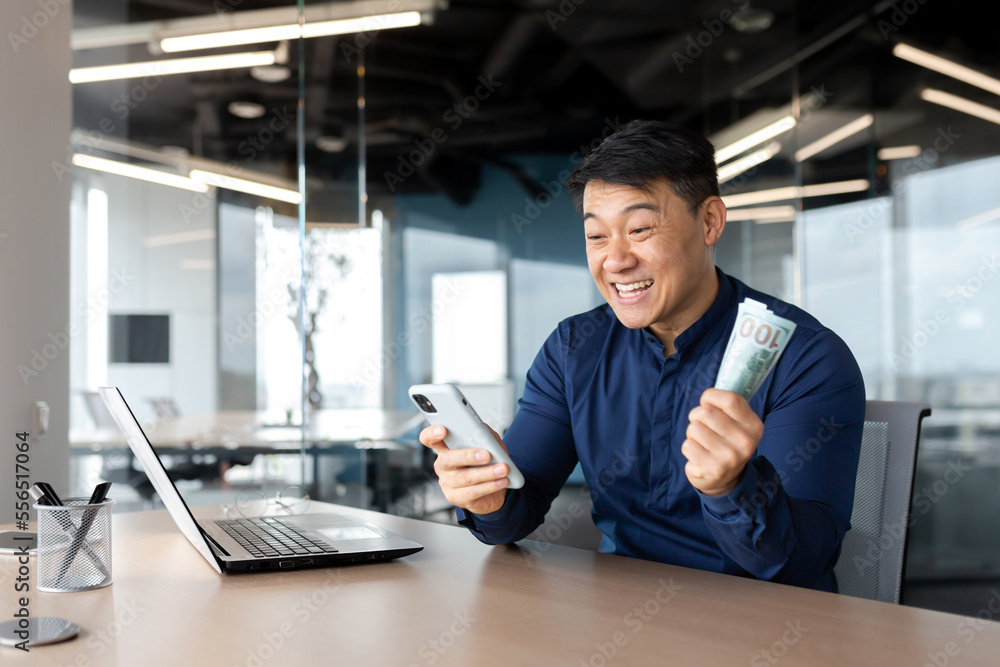 Happy young Asian man sitting in office holding cash money and phone. Rejoicing at the win, salary, placing bets online, celebrating.