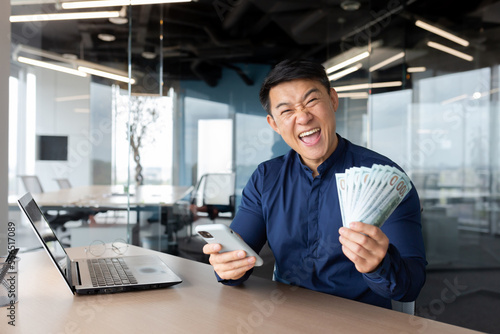 Happy young Asian man sitting in office holding cash money and phone. He looks at the camera, shouts with joy, winning, salary, places bets online, celebrates.