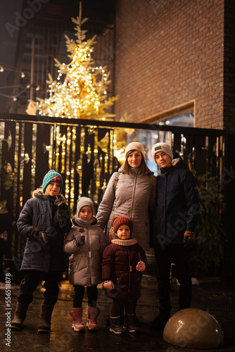 Mother with four kids stand against illuminated Christmas tree outdoor in evening.