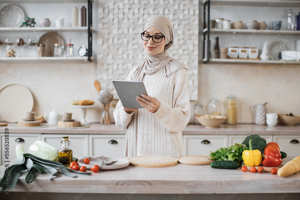 Cheerful young muslim woman in hijab cooking salad for dinner at home, looking at digital tablet, reading food blog, kitchen interior, copy space. Cooking, culinary concept. Cheerful young muslim woman in hijab cooking salad for dinner at home, looking at digital tablet, reading food blog, kitchen interior, copy space. Cooking, culinary concept.