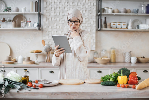 Cheerful young muslim woman in hijab cooking salad for dinner at home, looking at digital tablet, reading food blog, kitchen interior, copy space. Cooking, culinary concept.