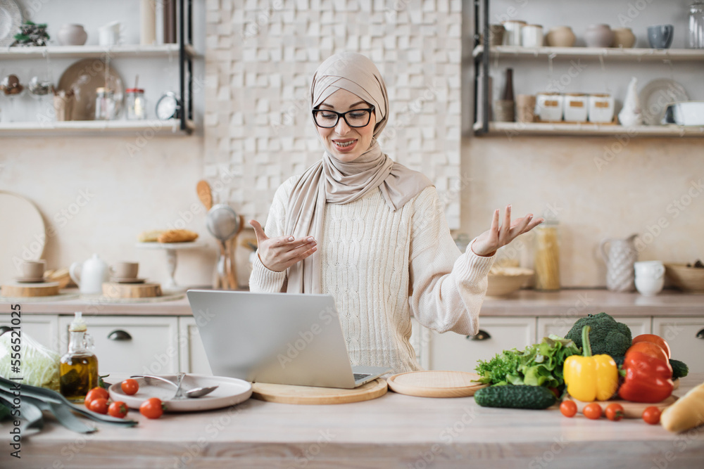 Muslim woman cooking delicious and healthy food in modern kitchen looking at laptop, while having online class, lesson or having video call with followers in social networks, media in internet at home Muslim woman cooking delicious and healthy food in modern kitchen looking at laptop, while having online class, lesson or having video call with followers in social networks, media in internet at home