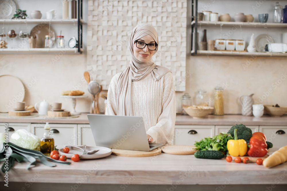 Cheerful young woman looking at an online recipe on a digital laptop pc. Attractive muslim female surfing the internet for recipes while making vegetarian salad in her bright kitchen. Cheerful young woman looking at an online recipe on a digital laptop pc. Attractive muslim female surfing the internet for recipes while making vegetarian salad in her bright kitchen.