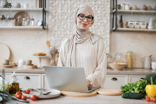 Cheerful young woman looking at an online recipe on a digital laptop pc. Attractive muslim female surfing the internet for recipes while making vegetarian salad in her bright kitchen.