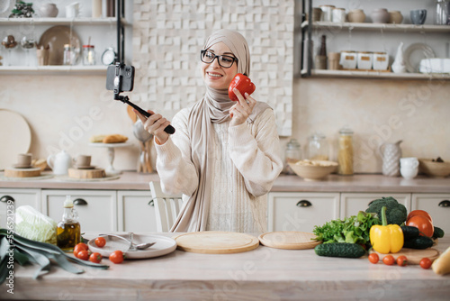 Happy young muslim woman using smartphone in selfie stick standing in kitchen in the morning. Happy female blogger holding pepper speaking with followers in social media about preparing healthy salad.