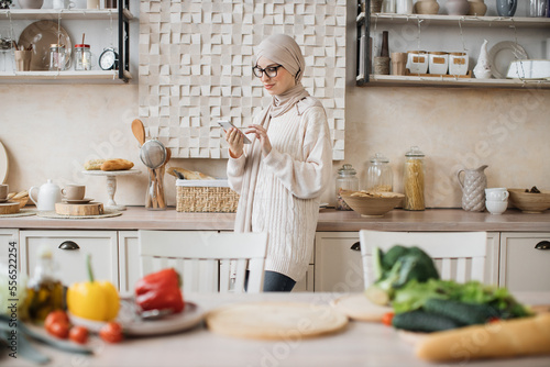 Attractive muslim woman preparing romantic dinner on home kitchen. Young female looking for good recipe at smart phone. Healthy food menu app. Cookbook mobile application. Technology and home