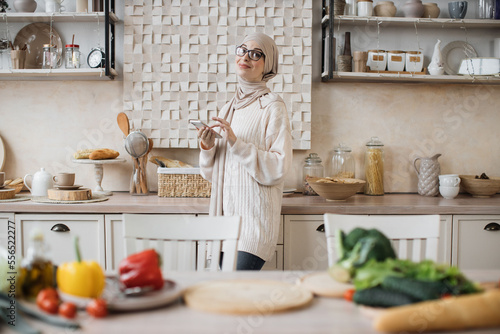 Happy young muslim woman using smartphone standing in kitchen in the morning. Pretty female in hijab reading news and enjoying leisure time at home, empty copy space.