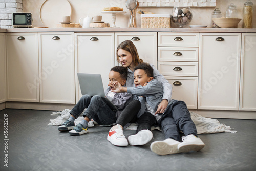 Happy multinational family of mother and two sons sitting on warm floor in light kitchen during weekend, resting and ordering food from a grocery store using laptop computer.