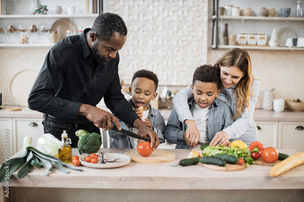 Happy multinational parents and two preschool sons preparing tasty food at kitchen. Mother and father teaching lovely kids to cook. Young mom and dad with their children spending time together at home