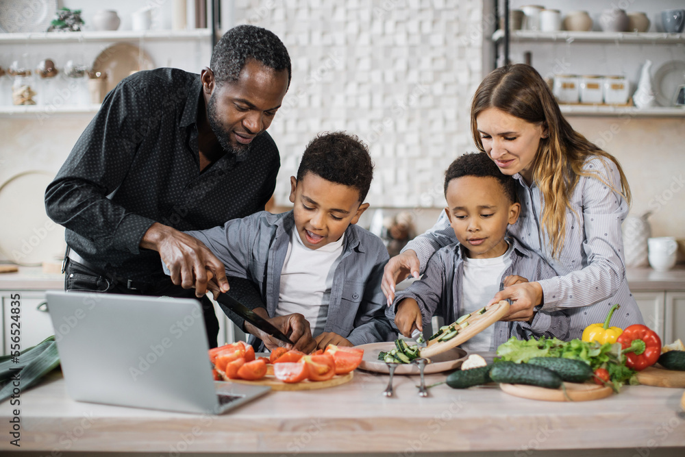 Happy african cute little sons and their young parents having online cooking class on laptop together using fresh vegetables and making salad, food in kitchen at home. Happy african cute little sons and their young parents having online cooking class on laptop together using fresh vegetables and making salad, food in kitchen at home.