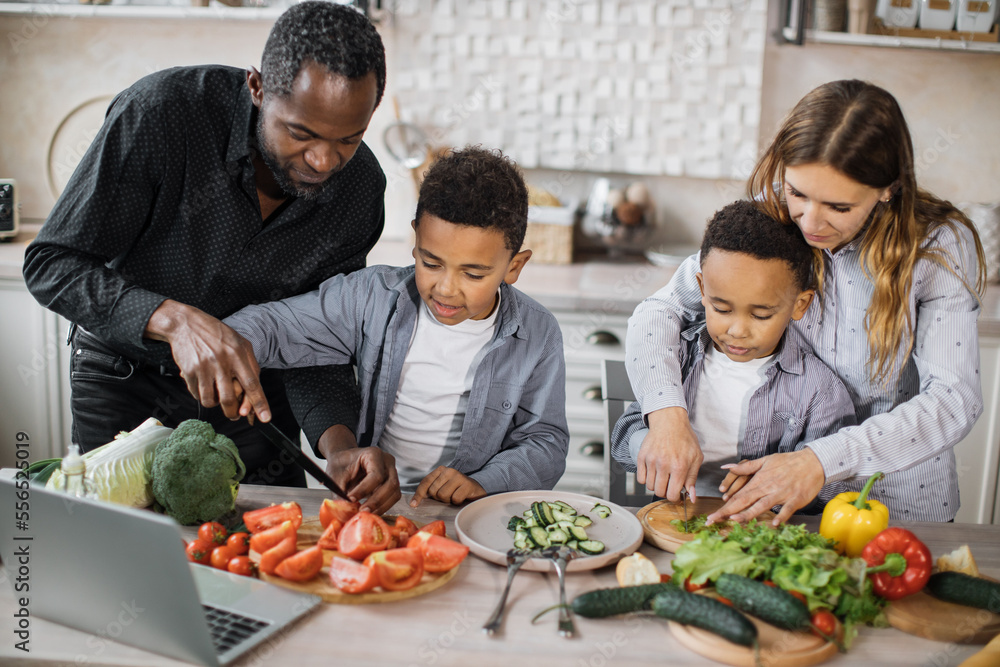 Happy young parents teaching little preschooler boys to chop vegetables with knife preparing salad for lunch together loving mom, dad and small sons kids cooking dinner together. Happy young parents teaching little preschooler boys to chop vegetables with knife preparing salad for lunch together loving mom, dad and small sons kids cooking dinner together.