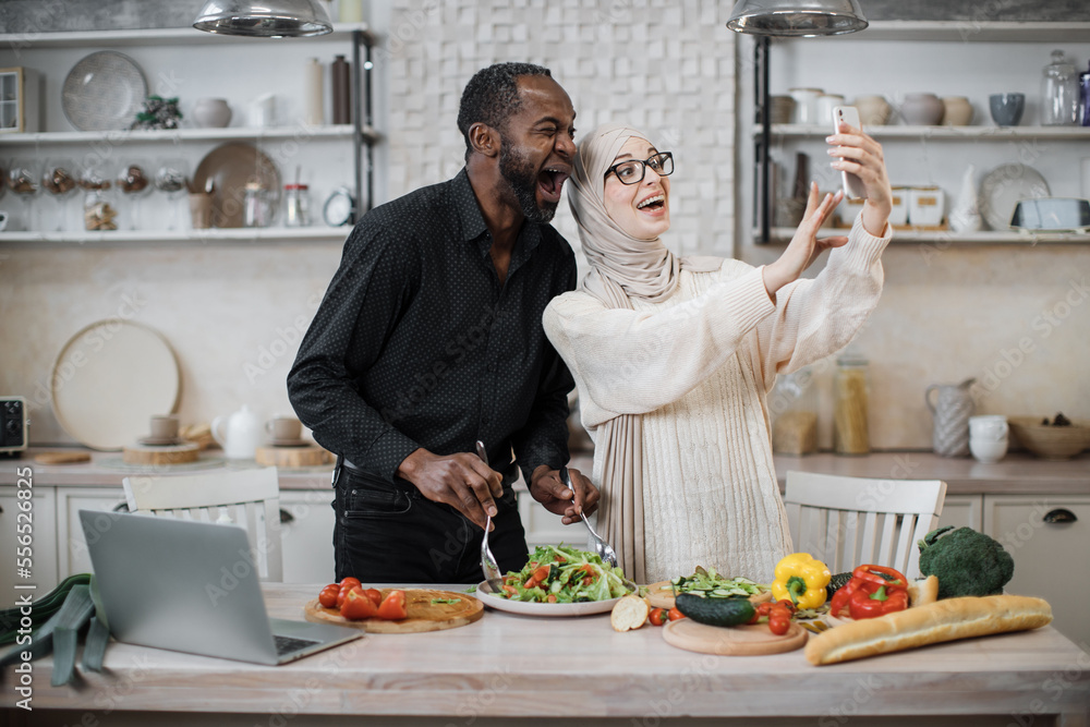 Cute multinational couple taking selfie or having video conversation with friends while having healthy breakfast at home, using brand new mobile phone, kitchen interior, copy space